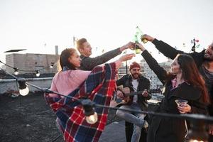 For the happiness. Group of young people having celebration at a rooftop with some alcohol and guitar playing