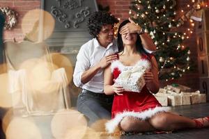 Beautiful moment. Man making surprise for his girlfriend. Gives New year present. Young couple sits on the floor of room of holiday decorated room photo
