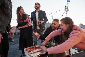 chico que se pone de pie le cuenta chistes a la hermosa chica morena. comiendo pizza en la fiesta de la azotea. buenos amigos tienen fin de semana con comida deliciosa y alcohol foto