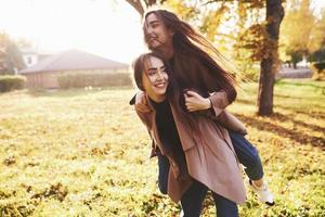 Young smiling brunette twin sisters having fun and doing a piggy back riding in autumn sunny park on blurry background