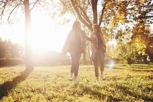 Young beautiful brunette twin girls walking together and holding hands, while looking at each other in autumn sunny park on blurry background photo