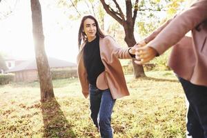 Young, smiling, pretty brunette girl is holding someone's hand while walking in autumn sunny park on blurry background photo