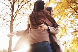 View from the back of young smiling brunette twin girls hugging in casual coat standing close to each other in autumn sunny park on blurry background photo