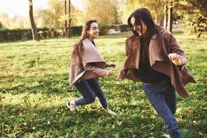 Side profile of young smiling brunette twin girls having fun, running and chasing each other in autumn sunny park on blurry background photo