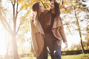 Bottom view of young smiling brunette twin girls hugging, looking at each other and walking in casual coat in autumn sunny park on blurry background photo