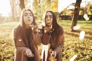 Young brunette twin sisters standing close to each other and blowing confetti into camera, holding some of those in their hands, wearing casual coat in autumn sunny park on blurry background photo