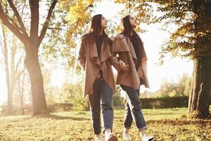 Profile view of young beautiful brunette twin girls walking with their hands in pockets close to each other and looking to a side together in autumn sunny park on blurry background