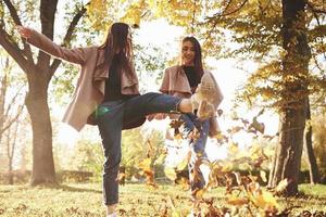Bottom view of young smiling brunette twin girls having fun and kicking leaves with their feet while walking in autumn sunny park on blurry background
