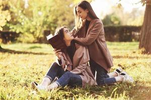 Brunette twin girls sitting on the grass and looking at each other. One of them is holding brown book, onother one is leaning on sisters shoulders from the back in autumn park on blurry background photo