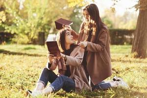 Brunette twin girls sitting on the grass with brown books in hands and looking at each other, when one of them is standing on her knees near the back of her sister in autumn park on blurry background photo