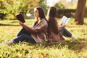 Side view of young pretty brunette twin sisters sitting back to back on the grass with legs slightly bent in knees reading brown books and wearing casual coat in autumn sunny park on blurry background photo
