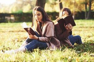 Young pretty brunette twin sisters sitting back to back on the grass with legs slightly bent in knees with brown books in hands, wearing casual coat in autumn sunny park on blurry background photo