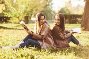Smiling brunette twin girls sitting back to back on the grass and looking at each other, legs slightly bent in knees, with brown books in hands, wearing casual coat in autumn park on blurry background photo