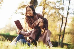 Young pretty brunette twin girls sitting on the grass. One of them is trying to read a brown books, while onother one is covering sisters eyes with her hands in autumn sunny park on blurry background photo