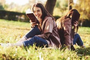 Laughing brunette twin girl sitting back to back on the grass with her sister, who is covering face with brown book, wearing casual coat in autumn sunny park on blurry background