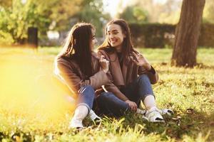 Young smiling brunette twin girls sitting on grass with legs crossed and slightly bent in knees wearing casual coat, chatting, looking at each other at autumn sunny park on blurry background