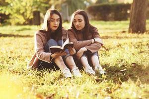 Young pretty brunette twin girls sitting on the grass with legs slightly bent in knees and reading in a brown book, wearing casual coat in autumn sunny parkr on blurry background