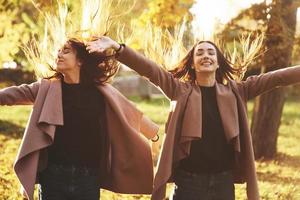 Young happy, smiling brunette twin girls standing and spreading hands wide open, their hair is dispelled up, wearing casual coat at autumn sunny park on the light blurry background. Freedom concept photo