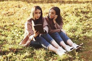 Young pretty brunette twin girls sitting on the grass with legs slightly bent in knees and looking in a brown book, wearing casual coat in autumn sunny weather on blurry background photo