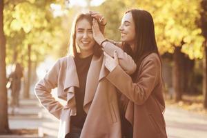 Young smiling brunette twin girls having fun with hands close to the head in one of them, in casual coat standing close at autumn sunny park alley on blurry background photo