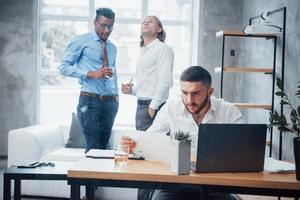 Employees at background. Young businessman sits on the chair in front of laptop and works with reports photo