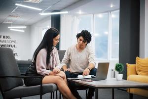 Sits on a grey chairs. Curly guy and brunette girl discusses the details of the contract in modern office photo