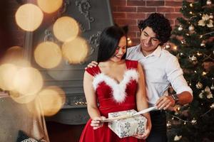 Opening the present. Beautiful couple celebrating New year in the decorated room with Christmas tree and fireplace behind