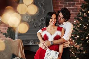 Cute hugs. Beautiful couple celebrating New year in the decorated room with Christmas tree and fireplace behind