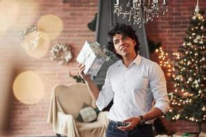 Tries to hear what's inside by shaking the present. Young man standing in the beautiful decorated room and holding white gift box in the New year time photo