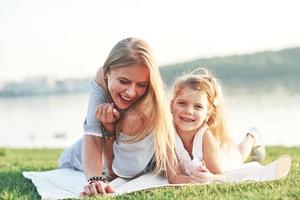 Smiling at the camera. Photo of young mother and her daughter having good time on the green grass with lake at background