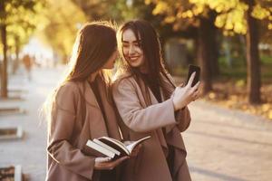 Jóvenes gemelas morenas sonrientes mirándose y tomando selfie con teléfono negro, mientras que una de ellas sostiene libros, usa abrigo, de pie en el callejón del parque soleado de otoño sobre fondo borroso foto