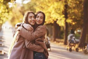 Side profile of young smiling brunette twin girls hugging and having fun in casual coat standing close to each other at autumn sunny park alley on blurry background photo