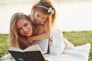 capta una atención muy especial. Mamá e hija leyendo un libro en un día soleado tendido en el césped con el lago al fondo foto