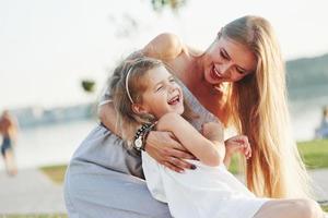 Everybody's laughing. Photo of young mother and her daughter having good time on the green grass with lake at background