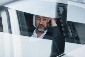 Seems tired. Reflection on window. Having business call while sitting at the back of modern luxury car photo