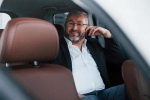 Cheerful senior businessman in eyeglasses sitting on the back of the car and smiling photo