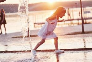 Running through the water. Young girl play in the fountain at the summer heat and lake and woods background