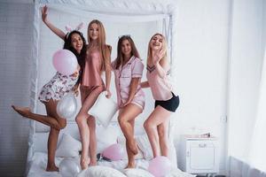Posing for the picture. Standing on the luxury white bad at holiday time with balloons and bunny ears. Four beautiful girls in night wear have party photo