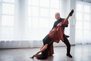 Girl is almost upside down. Young pretty woman in red and black clothes dancing with bald guy in the white room photo