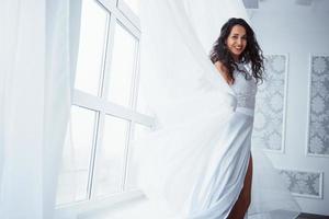 Sincere smile. Beautiful woman in white dress stands in white room with daylight through the windows