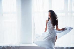 Horizontal photo. Beautiful woman in white dress stands in white room with daylight through the windows