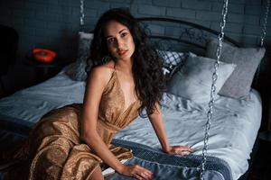 mujer elegante. Chica en vestido dorado se sienta en la cama blanca en las cadenas de apartamentos de lujo foto