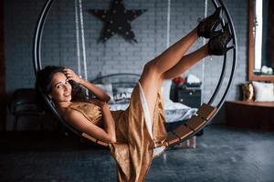 Pretty woman in golden colored dress sits on the stylish circle shaped bench on the chain. Luxury room photo