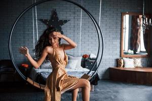 Motions with hand. Pretty woman in golden colored dress sits on the stylish circle shaped bench on the chain. Luxury room photo
