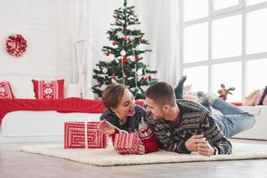 Encantadora pareja joven tumbado en la sala de estar con árbol de Navidad verde en el fondo foto