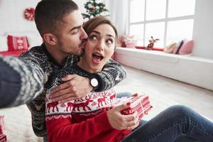 mujer camina hacia su novio mientras sostiene el regalo. Hombre tomando selfie de él y su esposa vestidos con ropa navideña y sentados en el piso de la hermosa habitación decorativa