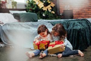 Friends hug each other. Christmas holidays with gifts for these two kids that sitting indoors in the nice room near the bed