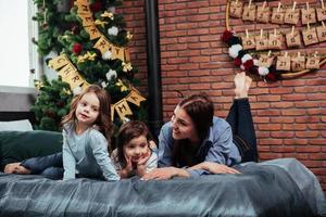 Smiling and having good time. Mother and two kids laying down on the bed with room that decorated with christmas tree