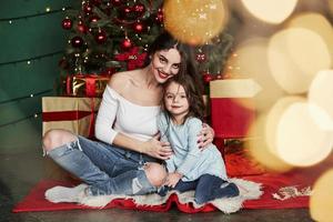 Holidays conception. Cheerful mother and daughter sitting near the Christmas tree that behind. Cute portrait photo