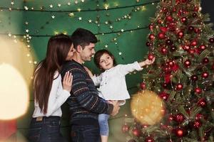 Cute little girl is touching red holiday sphere. Happy family celebrating. New year and standing near Christmas tree photo